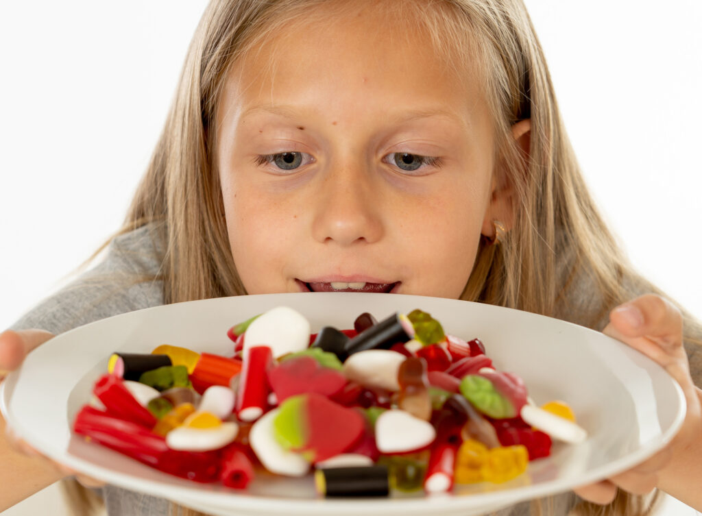 pretty happy sugar high caucasian female child eating dish full of candy holding fork and knife in sweet sugar abuse dangerous diet and unhealthy nutrition concept isolated on white background